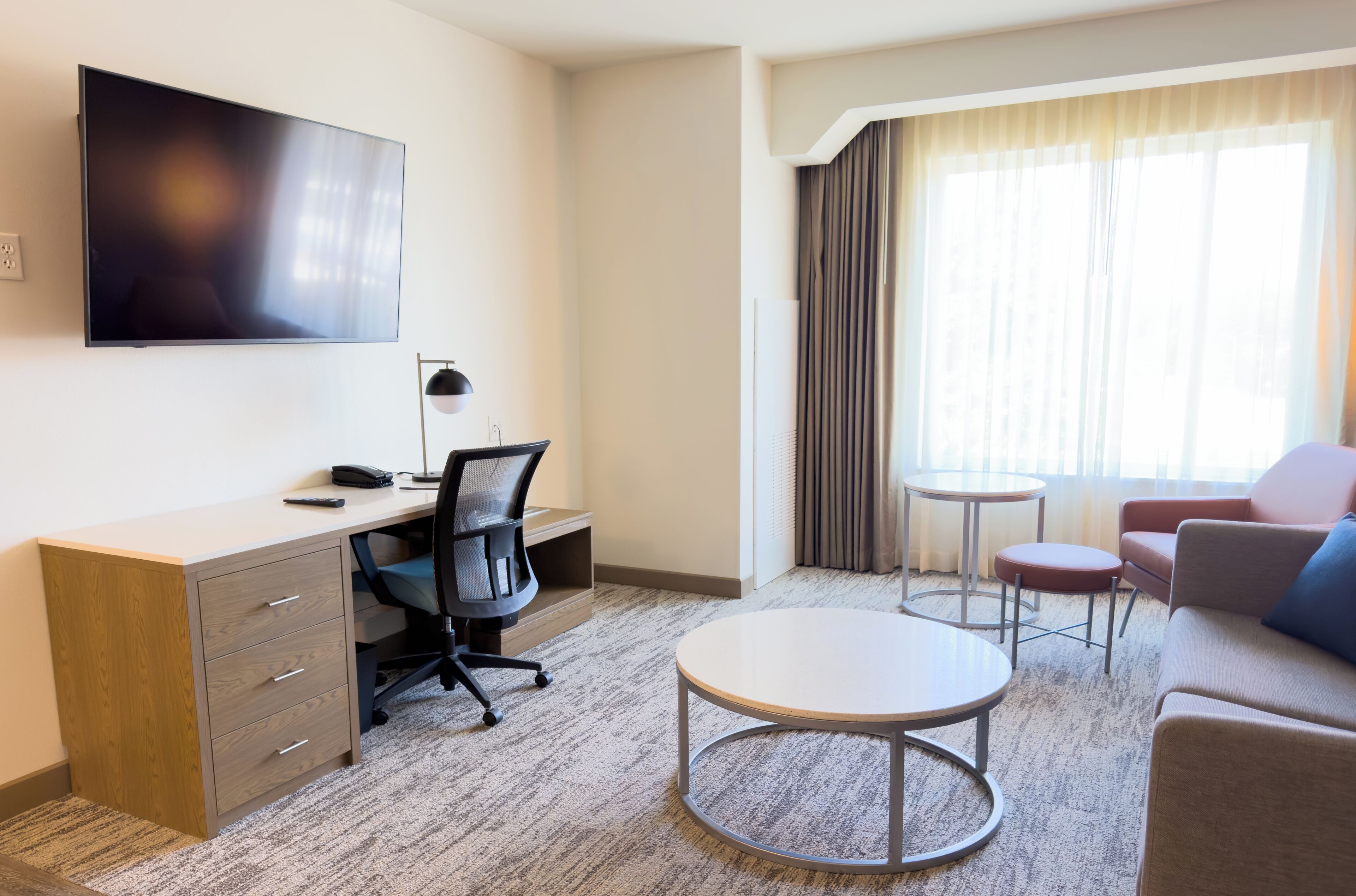A hotel room with a flat-screen TV, a desk with a chair, a round coffee table, a sofa, and a small table with two chairs.