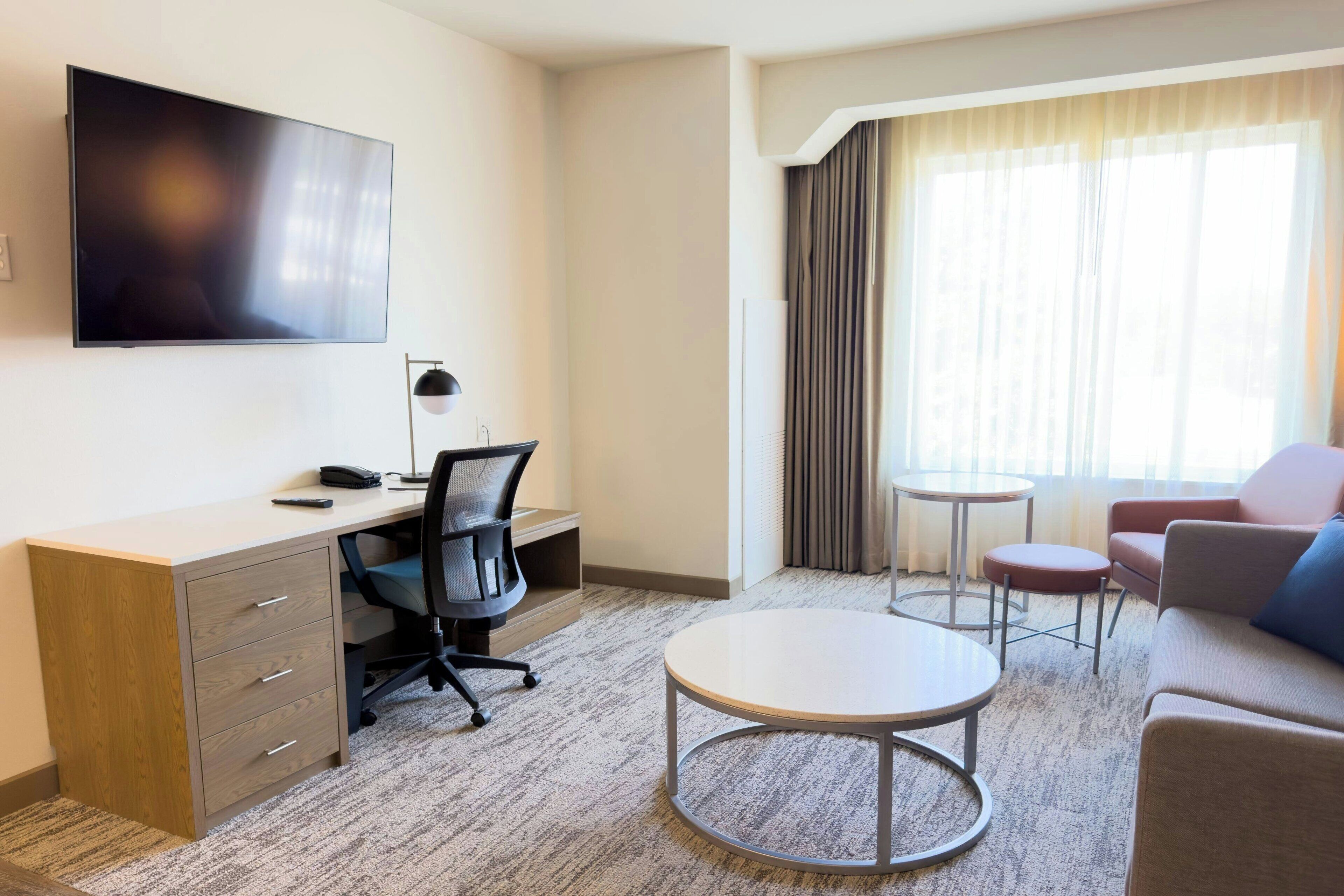 A hotel room with a desk, chair, TV, small round table, and sofa.