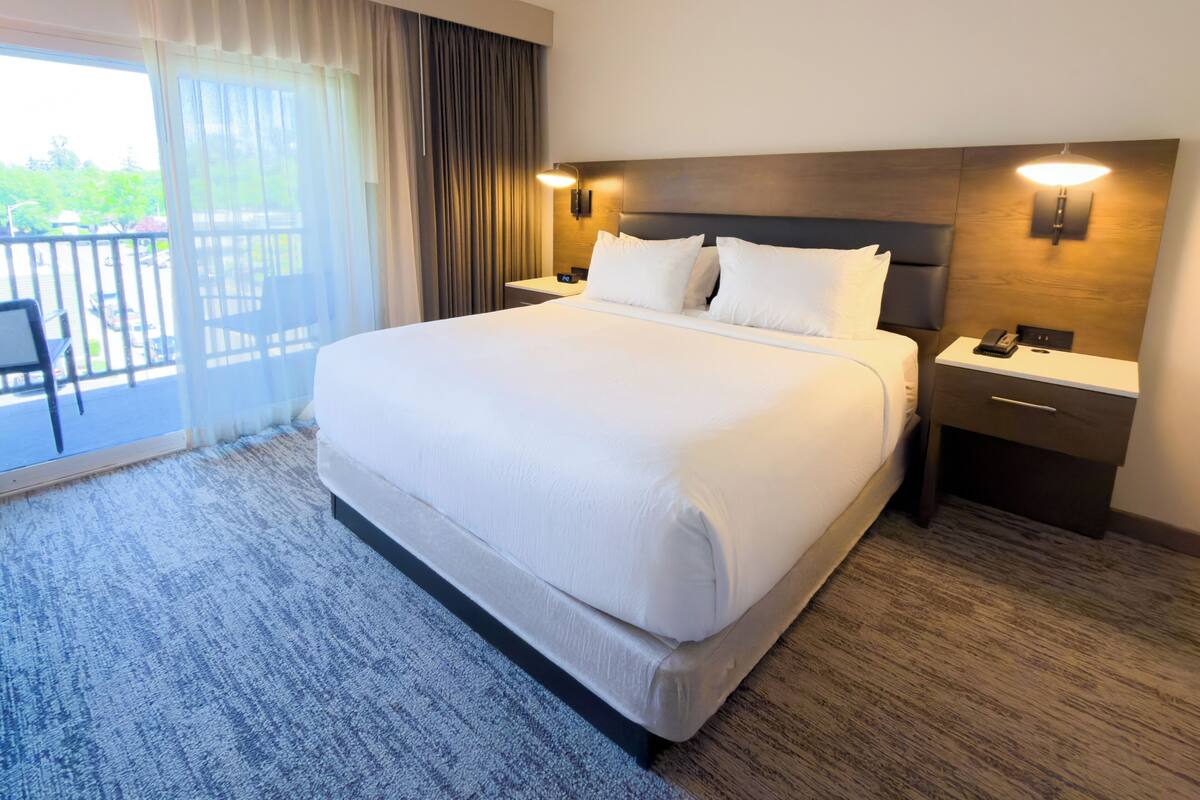 Quarto de hotel com cama grande, mesa de cabeceira, abajur e vista para o exterior.