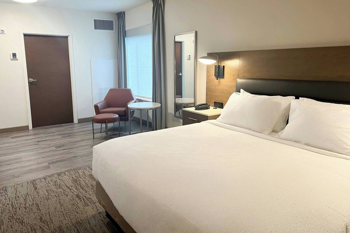 Quarto de hotel com uma cama grande, uma escrivaninha com cadeira, uma mesinha redonda, um espelho e uma porta.