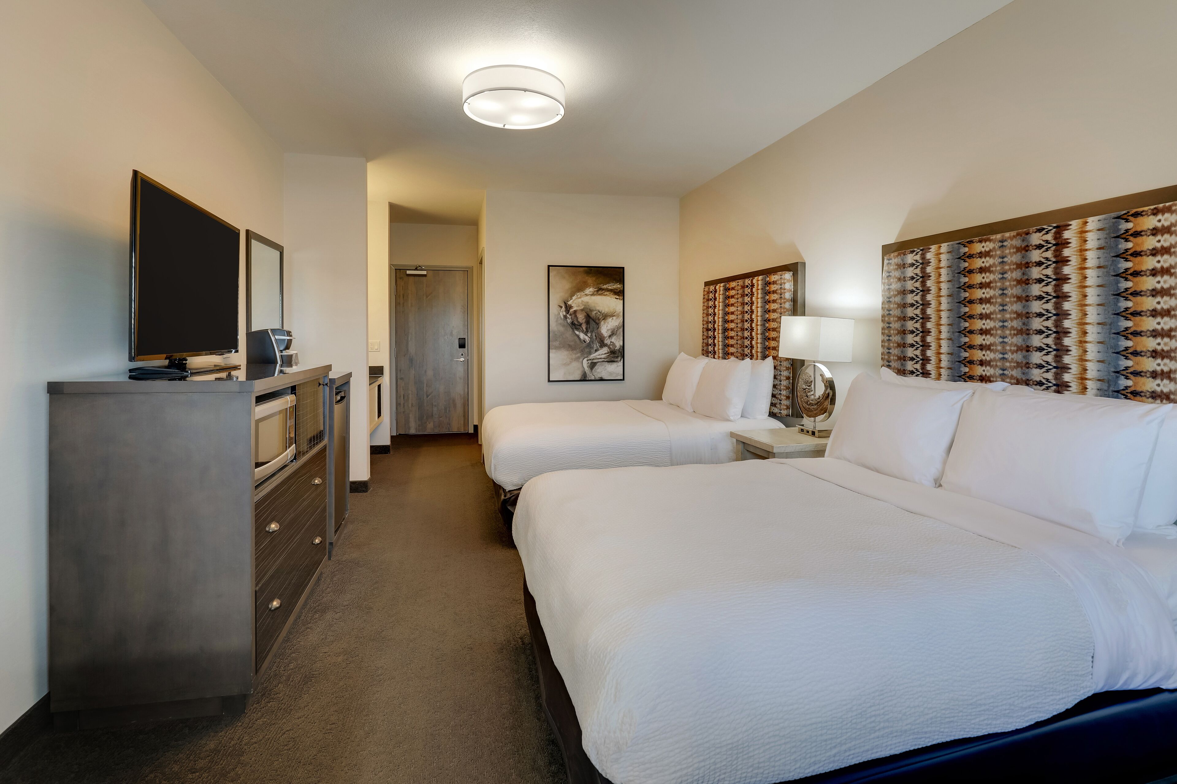 A hotel room with two beds, a desk with a flat-screen TV, a dresser, and a large patterned headboard.