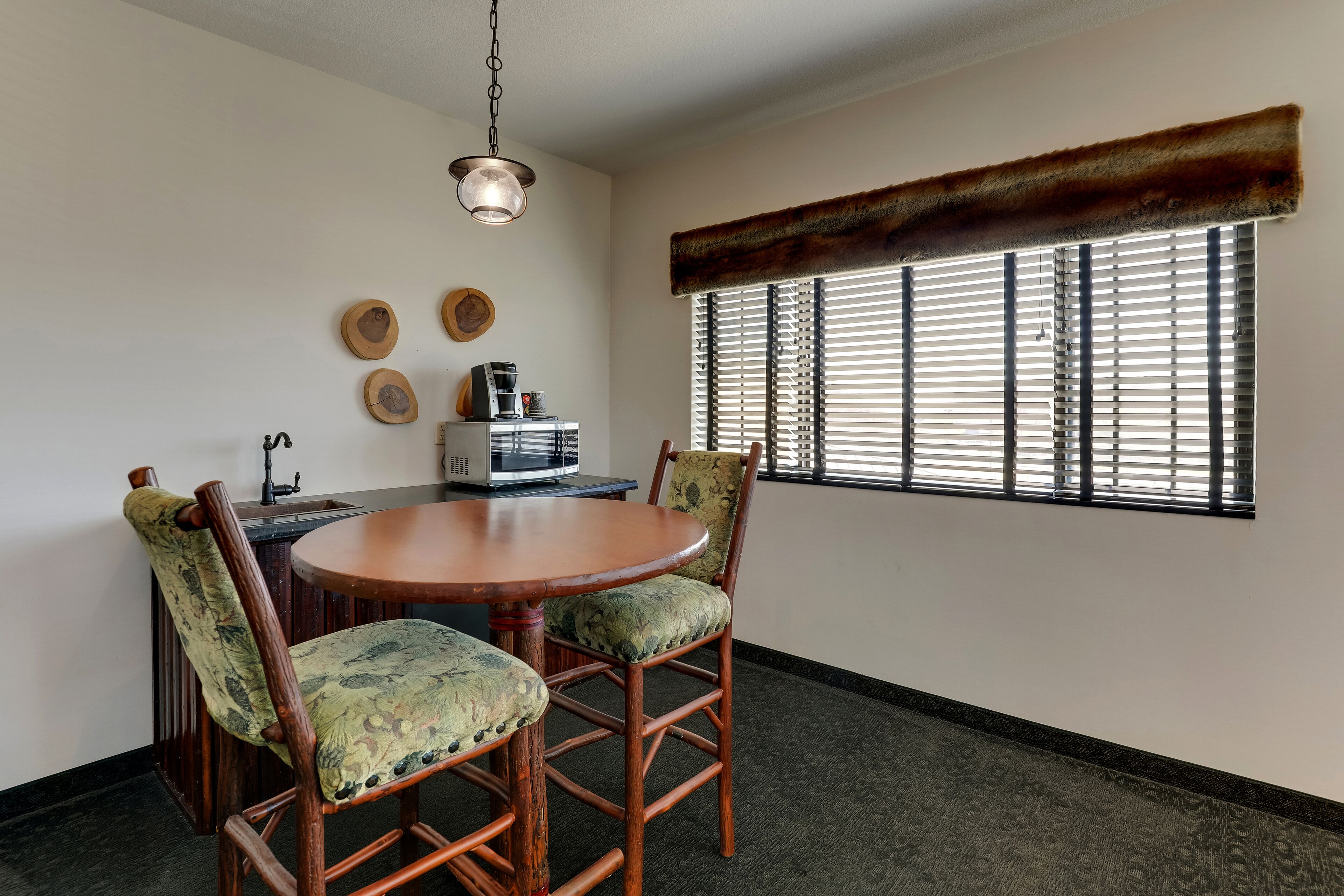 A dining area with a round table, two chairs, a coffee maker, and a window with blinds.