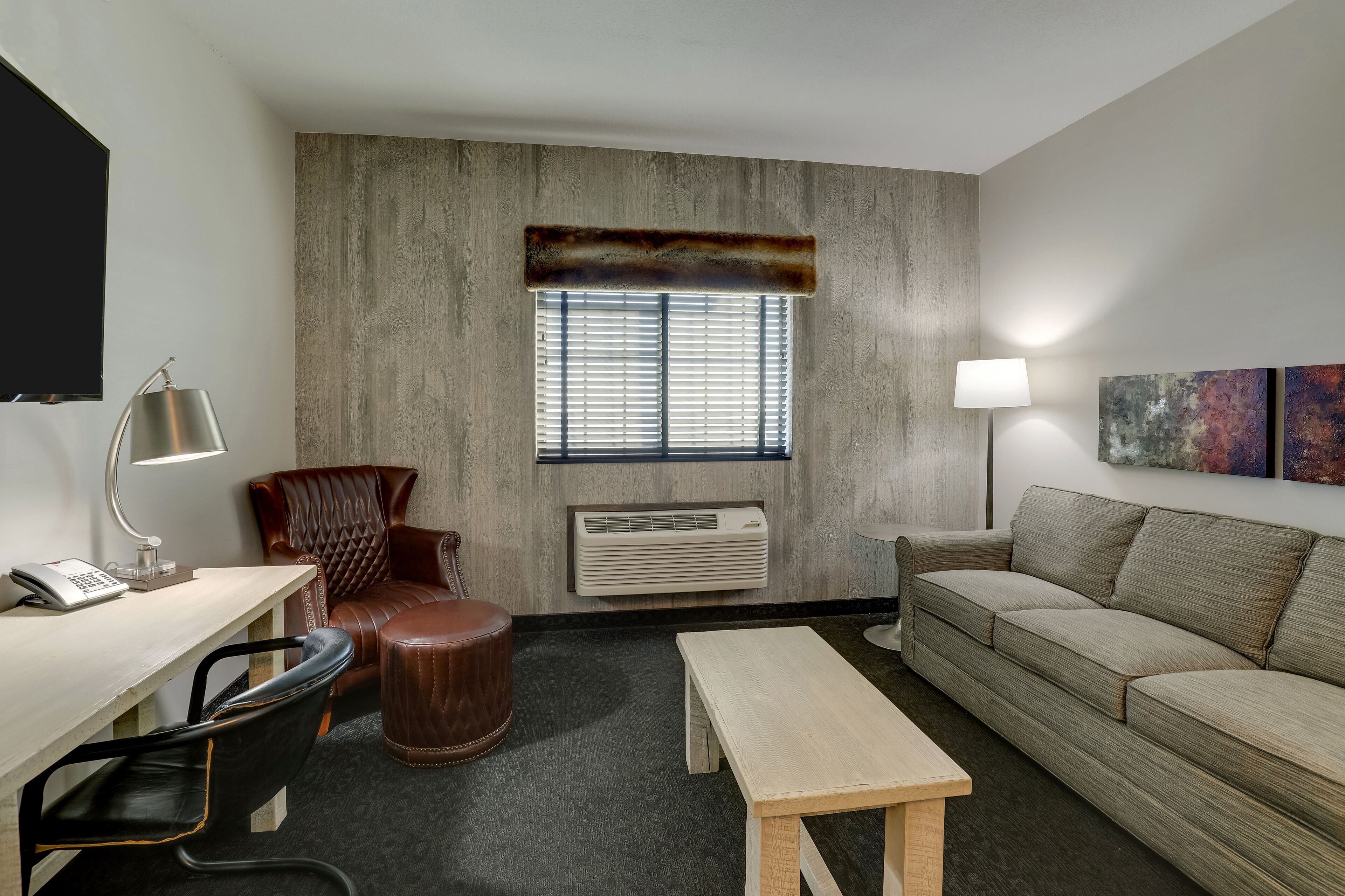 A hotel room with a desk, leather chair, sofa, and a window with blinds.