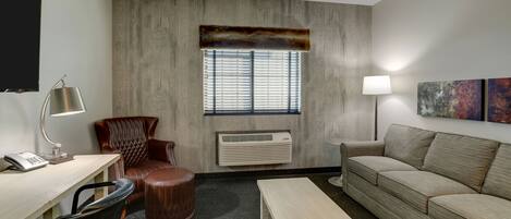 A hotel room with a desk, leather chair, sofa, and a window with blinds.