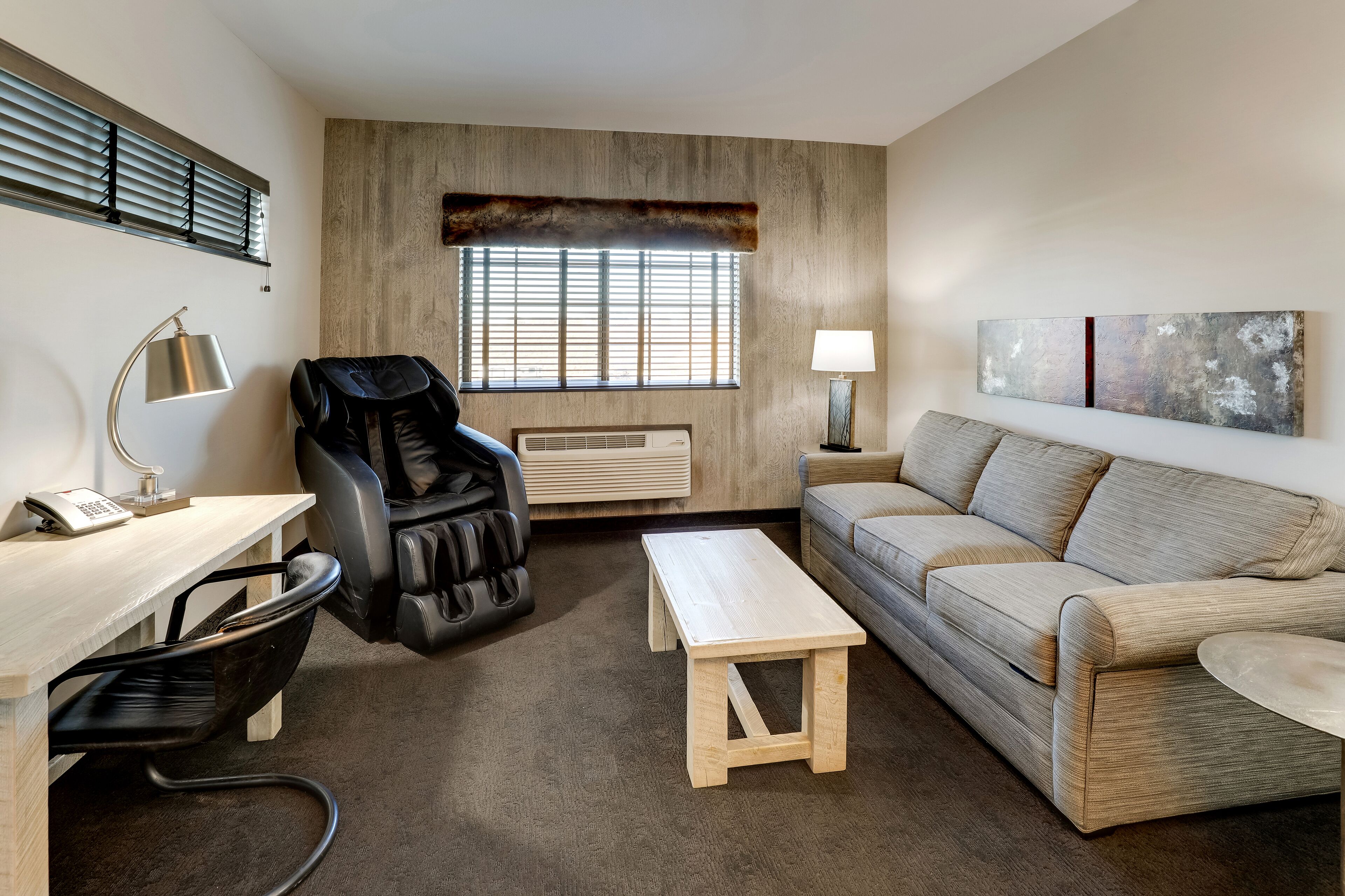Un salon moderne comprenant un canapé, un bureau avec une chaise, un fauteuil de massage, une petite table en bois et une fenêtre avec des stores.