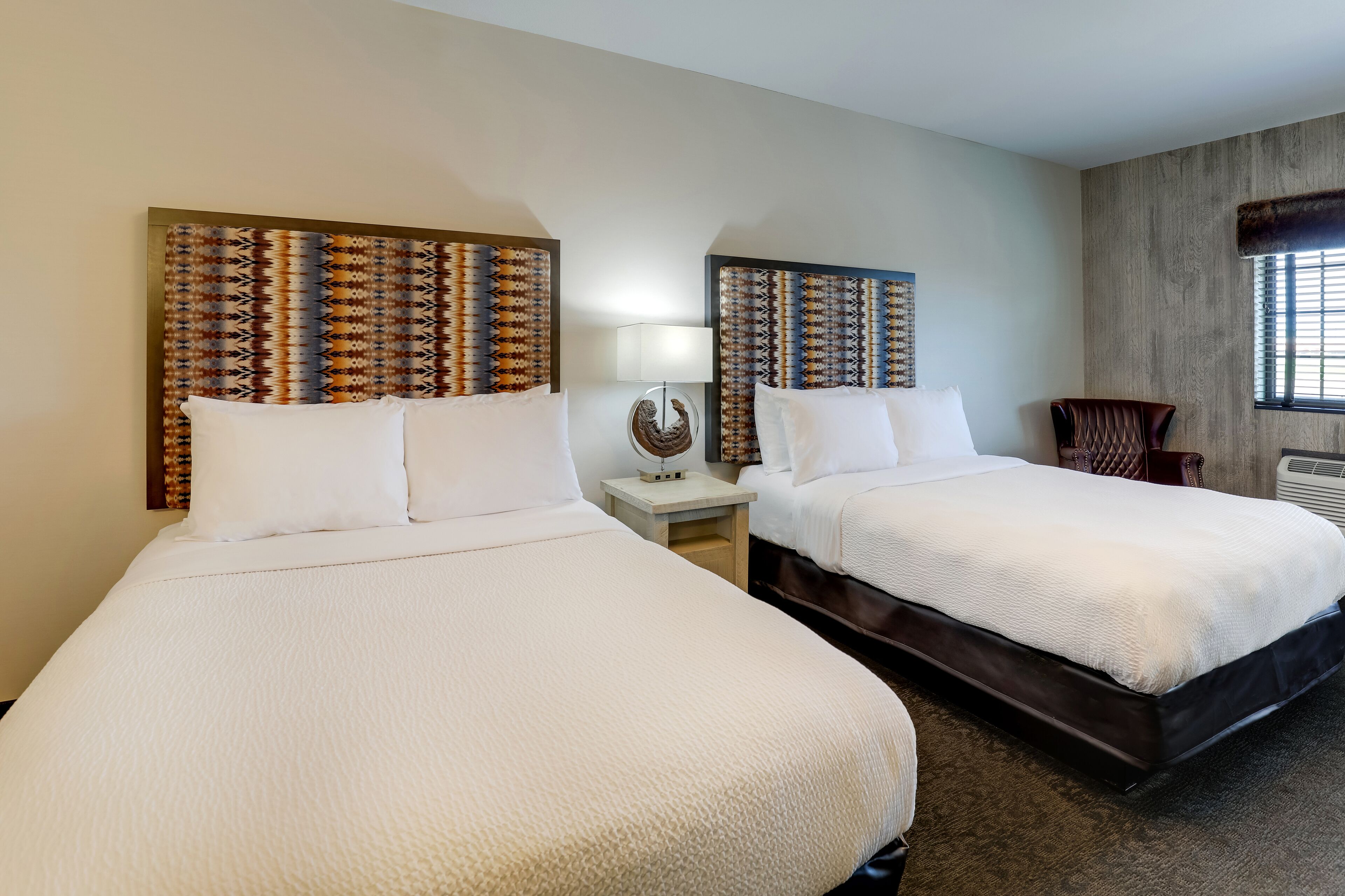 A hotel room with two beds, a nightstand with a lamp, a decorative wall piece, and a window with blinds.