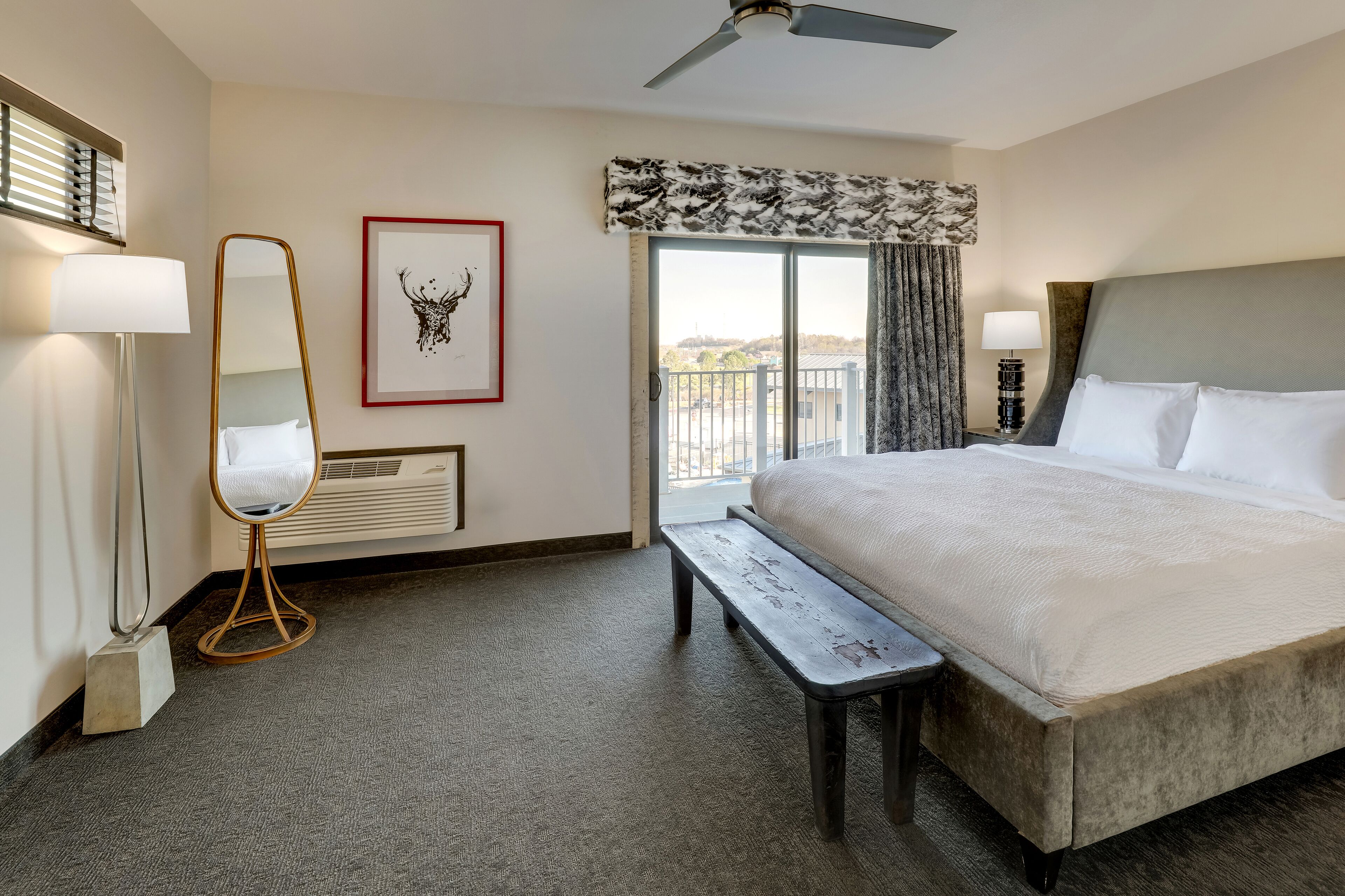 Une chambre d’hôtel avec un grand lit, un banc, une lampe sur pied, un miroir, un tableau encadré et une fenêtre donnant sur un beau paysage.