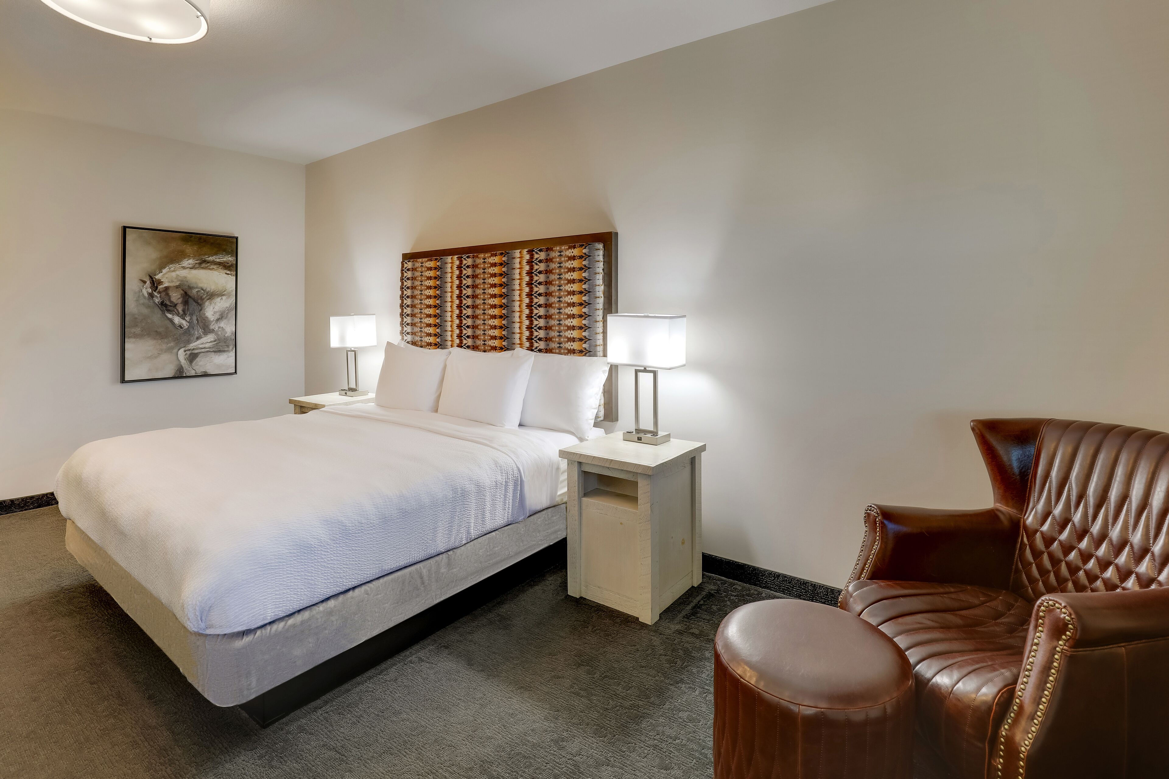 A hotel room with a bed, two bedside tables with lamps, a leather armchair, and a wall-mounted headboard.