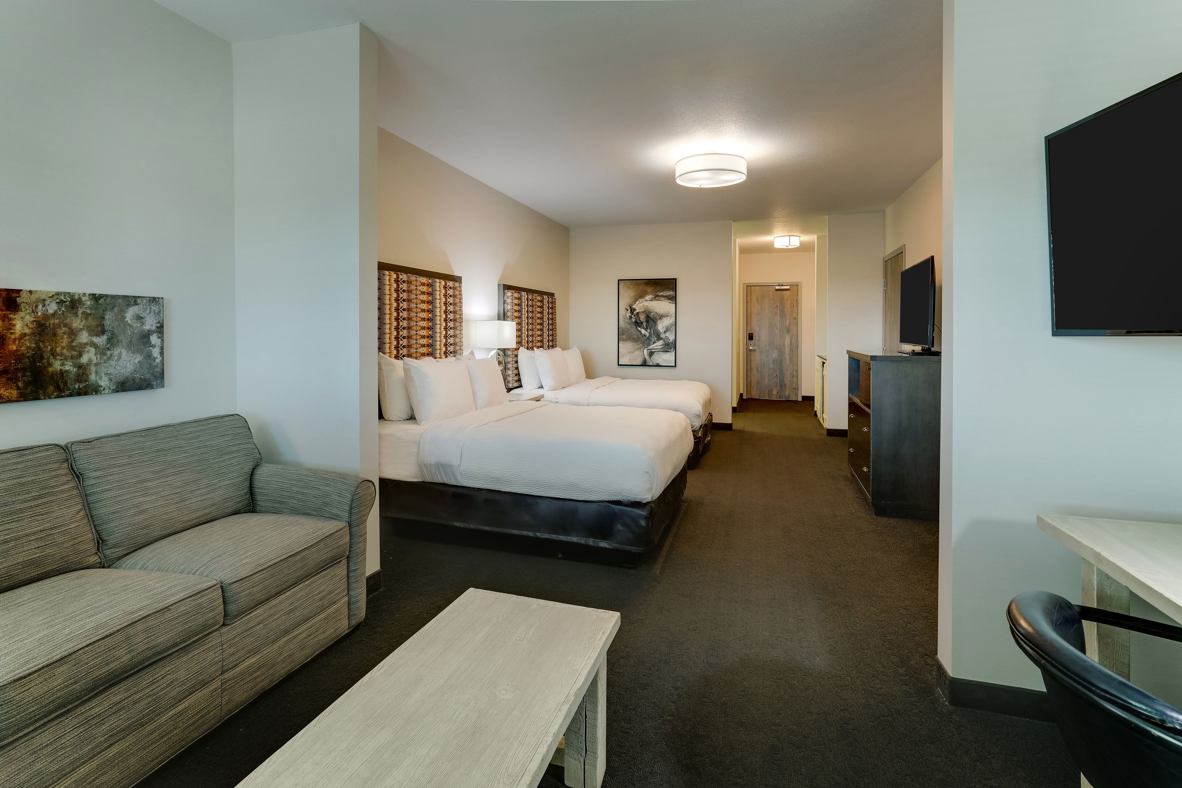 A hotel room with a sofa, two beds, a desk, a TV, and artwork on the walls.