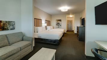 A hotel room with a sofa, two beds, a desk, a TV, and artwork on the walls.