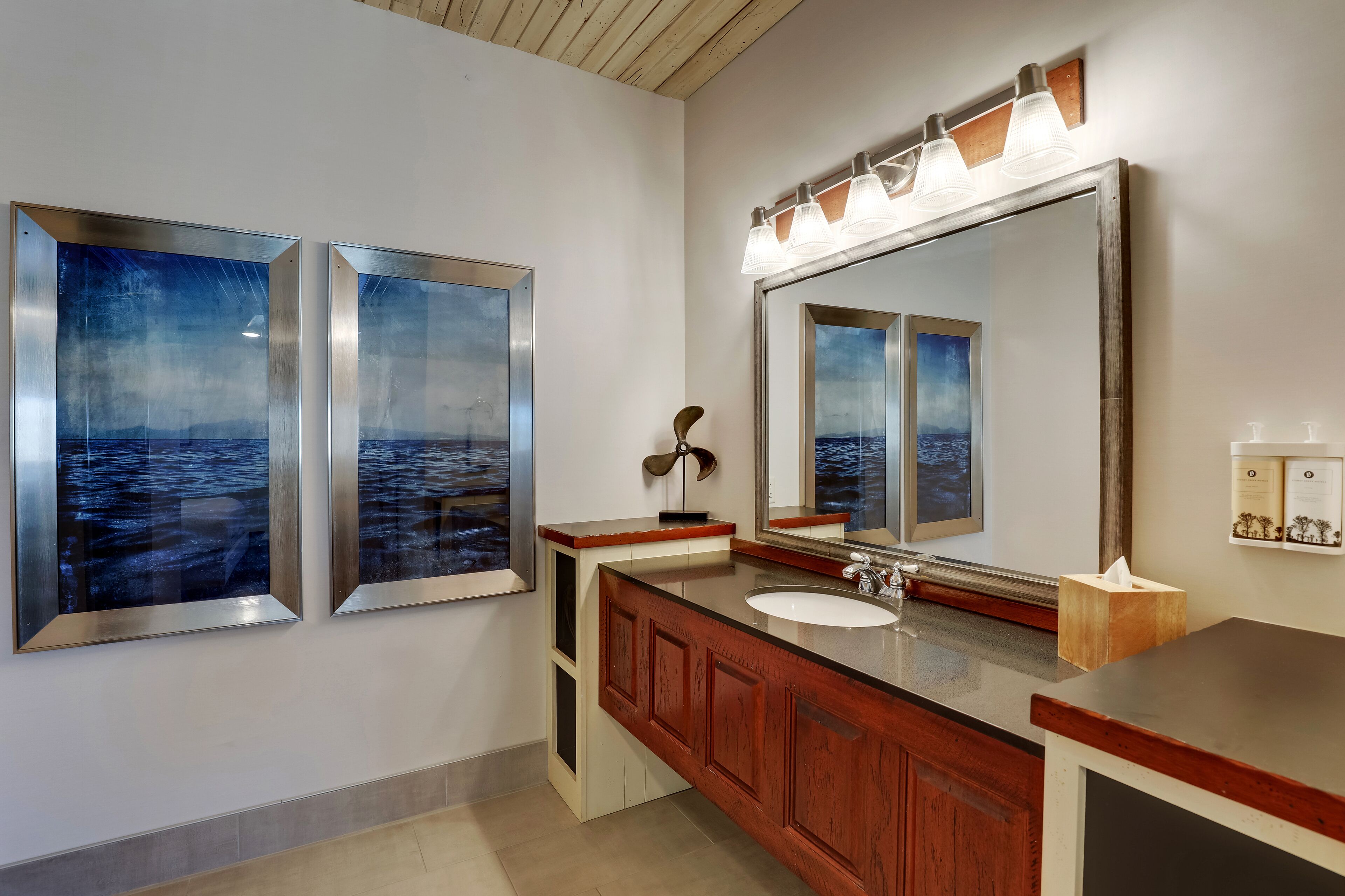 A bathroom with a large mirror, wooden vanity, and two framed artwork depicting seascapes.