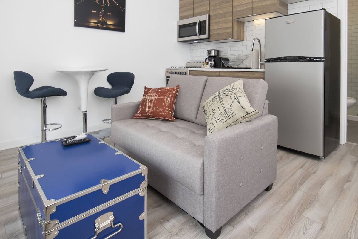 A compact living space with a kitchenette, a grey sofa, a blue suitcase, and a small dining table.