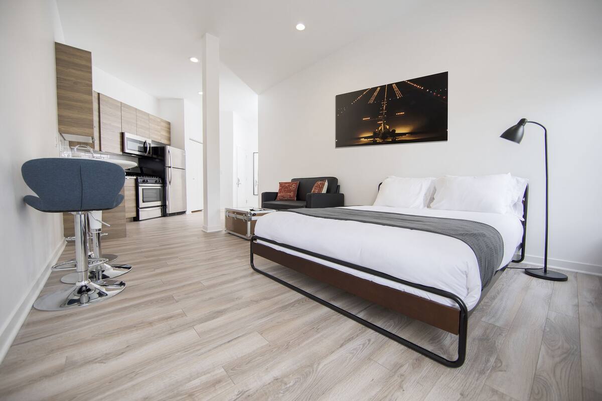 A modern bedroom with a bed, bedside table, lamp, and a view of the kitchen.