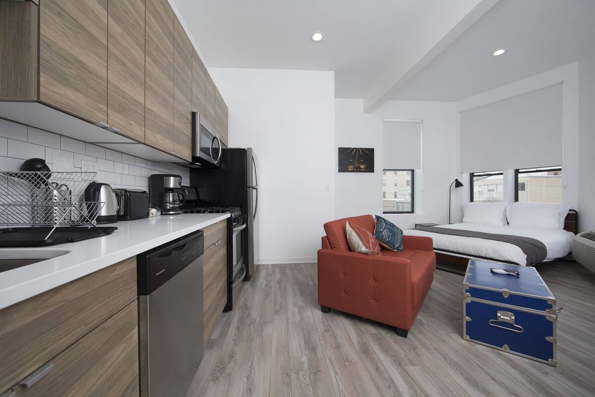 A modern kitchen with wooden cabinets, a microwave, and a coffee maker. There is a red sofa and a blue suitcase in the living area.