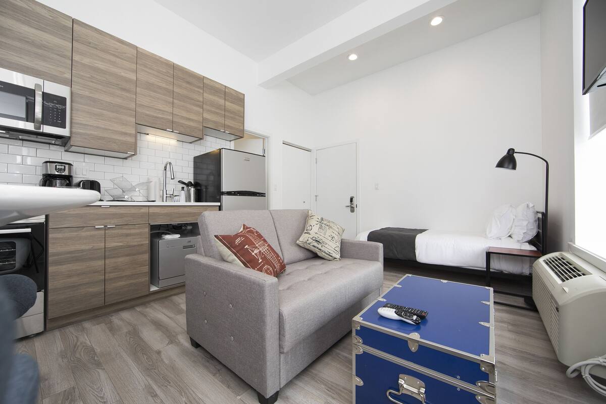 A modern kitchen with wooden cabinets, a microwave, and a refrigerator. There is a grey sofa with a remote control on a blue chest of drawers. A bed with white bedding is visible in the background.