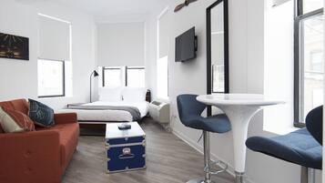 A modern hotel room with a bed, a sofa, a dining table, and a TV mounted on the wall.