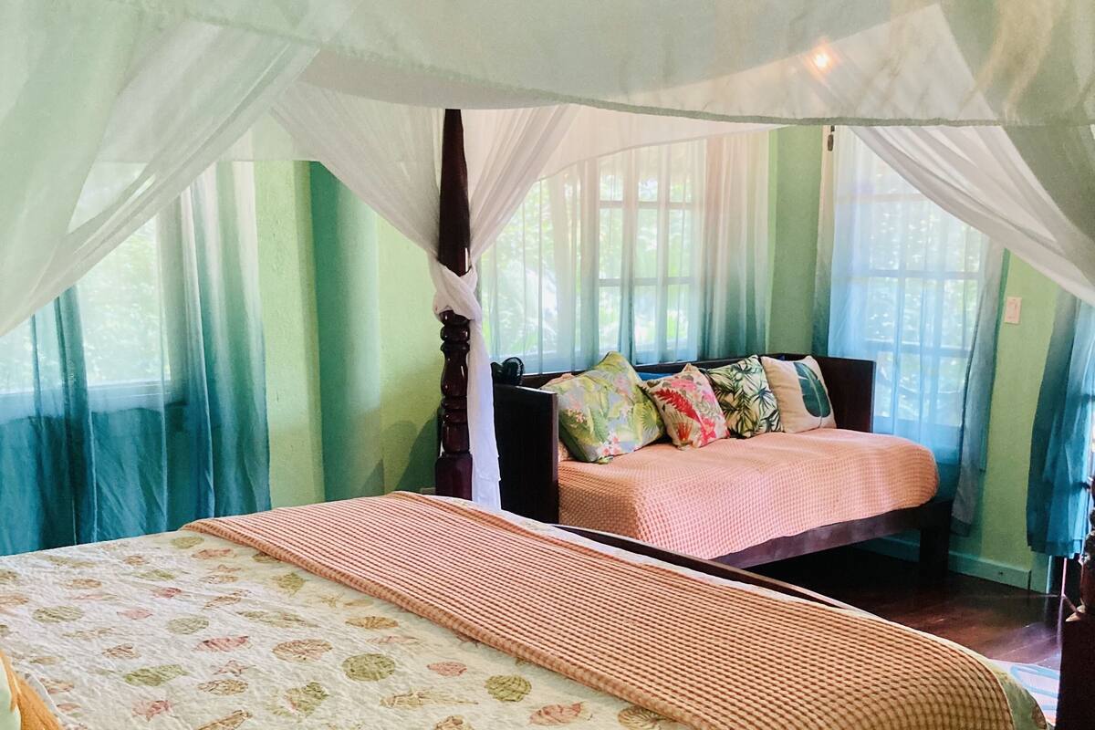 A bedroom with a canopy bed, a separate bed with a floral bedspread, and a view of the outdoors through large windows.