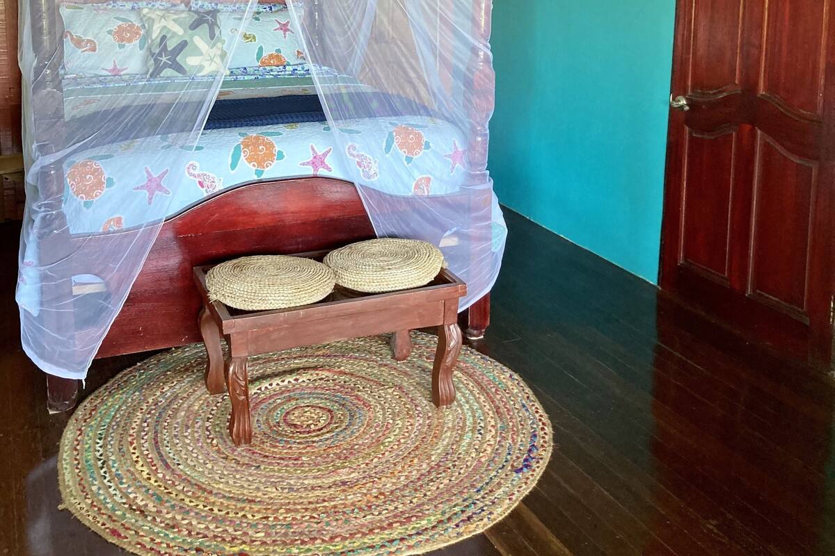 A room with a bed under a mosquito net, a wooden bench, a ceiling fan, and a door.
