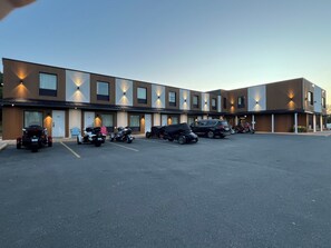 Een modern gebouw met meerdere wooneenheden, een parkeerplaats en verschillende voertuigen, waaronder motorfietsen en een auto.