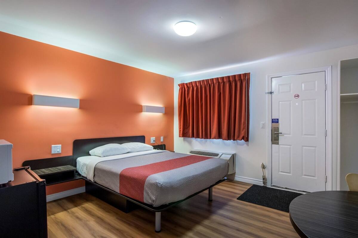 Quarto de hotel com uma cama, escrivaninha, cadeira, porta e janela com cortinas.