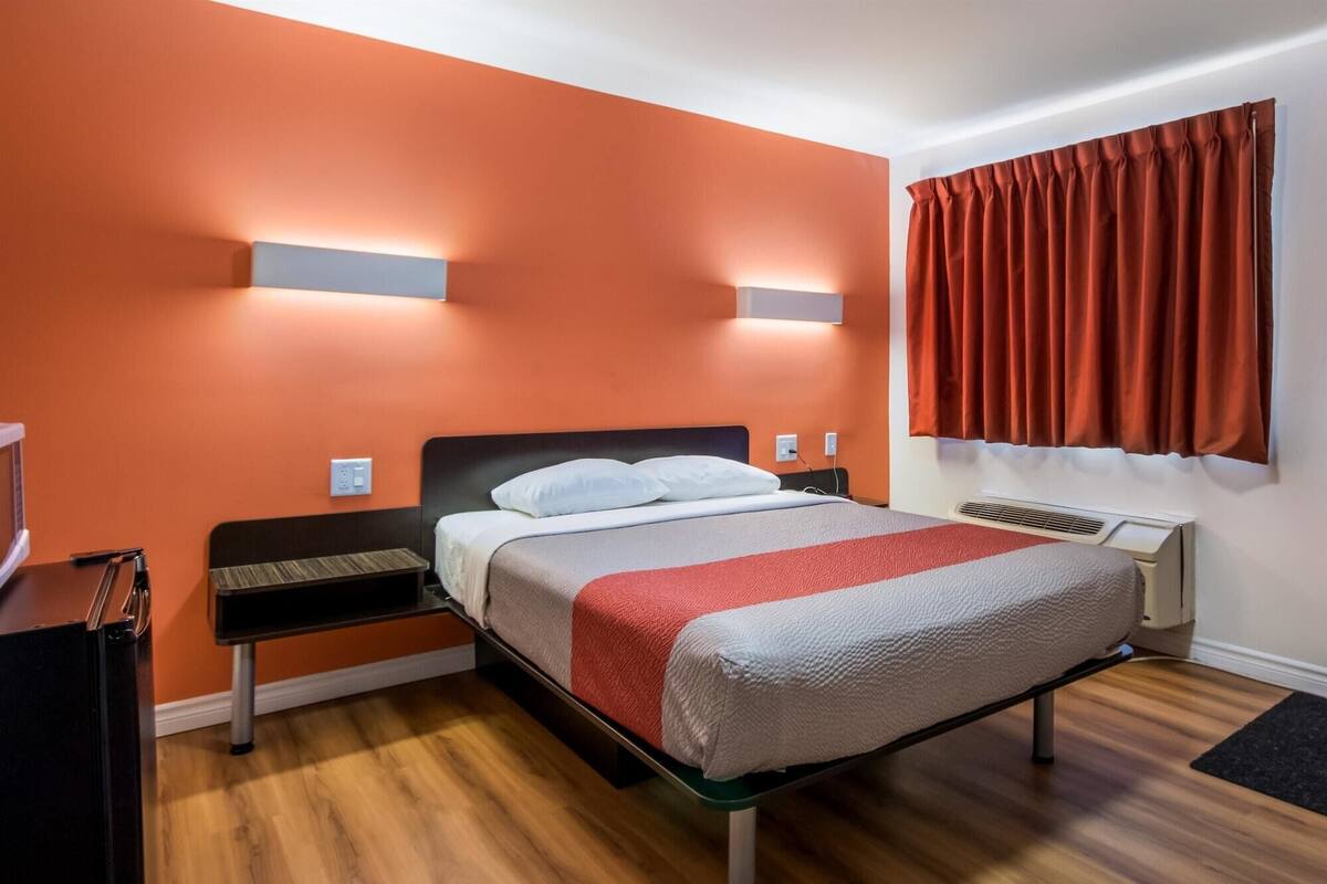 Quarto de hotel com uma cama de solteiro, mesa de cabeceira, televisão e janela com cortinas.