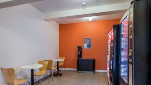 Ein kleiner, hell erleuchteter Raum mit orangefarbenen Wänden, einer Kaffeemaschine, einem Automaten und einigen Tischen und Stühlen.