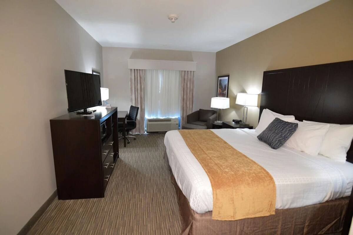 Ein Hotelzimmer mit einem großen Bett, einem Schreibtisch mit Fernseher, einem Sessel, einer Schreibtischlampe und einem Fenster mit Vorhängen.