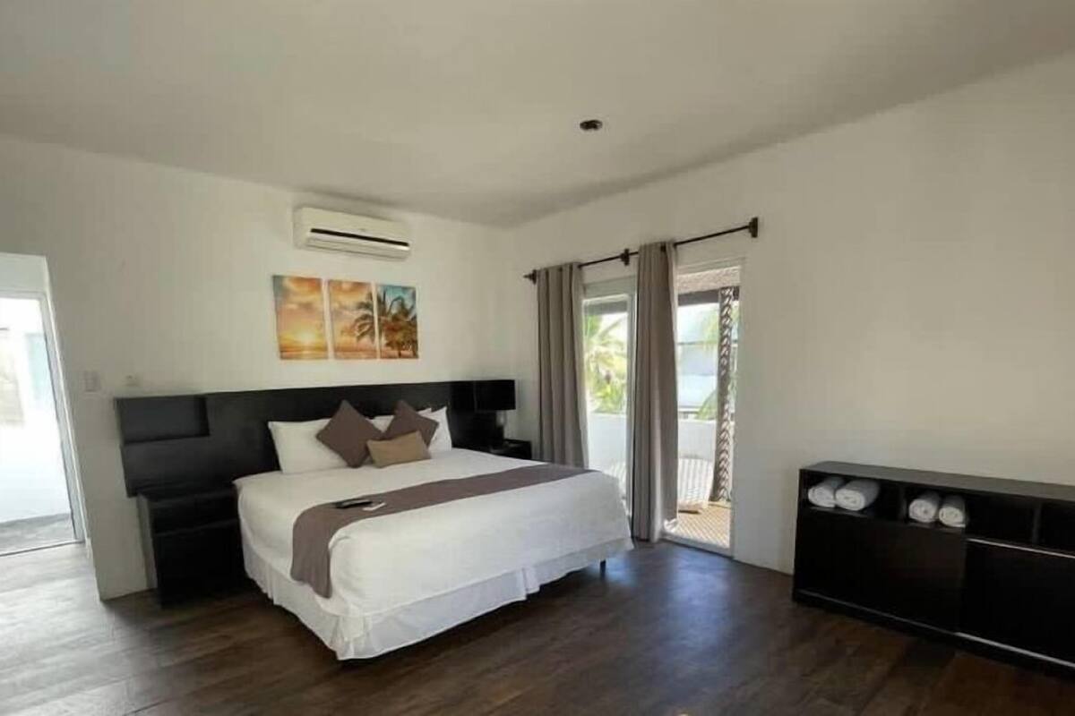 Ein Schlafzimmer mit einem großen Bett, einem dunklen Kopfteil, einem Nachttisch, einer Klimaanlage, einem an der Wand befestigten Fernseher und einer Glastür zu einem Balkon.
