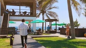 Een houten pad dat leidt naar een overdekt terras met tafels en stoelen, met uitzicht op de zee.