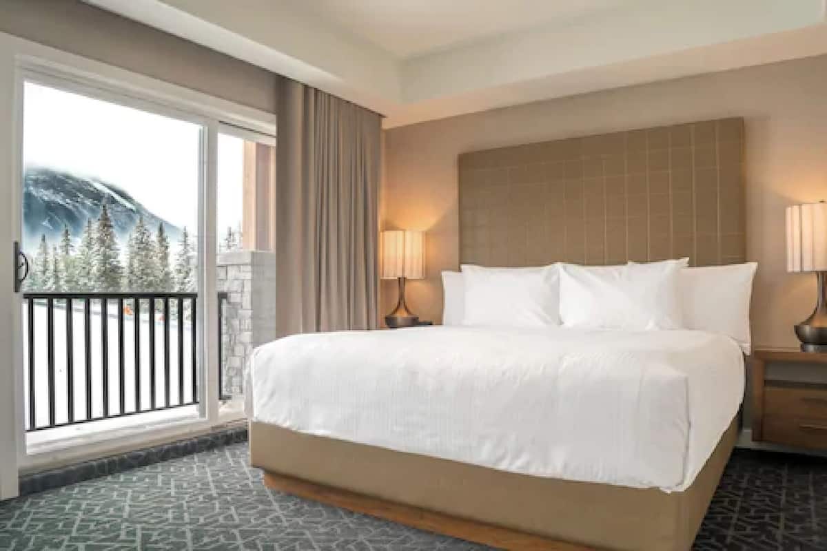 A hotel room with a large bed, two bedside lamps, a view of trees and mountains through a sliding glass door, and a balcony with a railing.