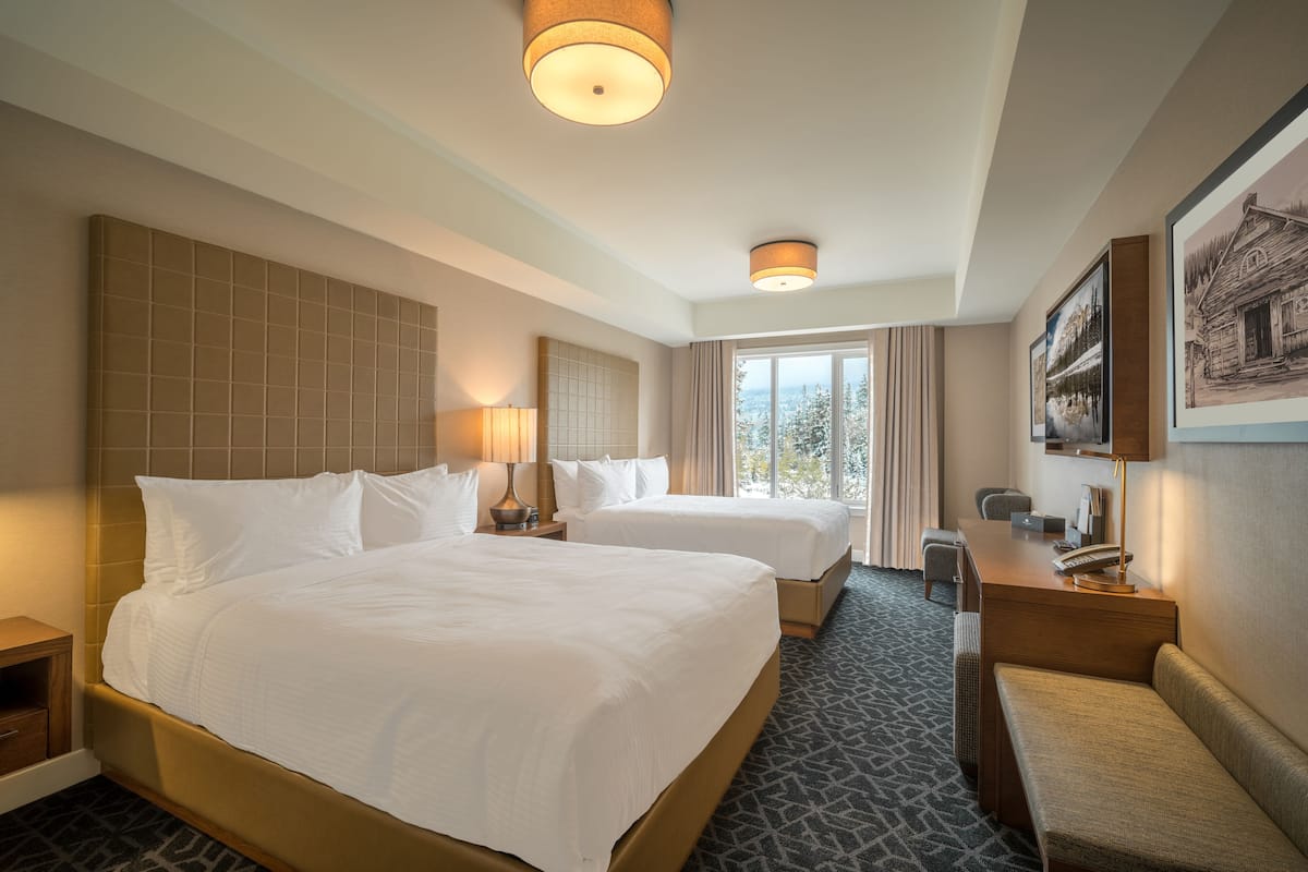 A hotel room with two beds, a desk, a chair, a bench, and a large window with a view.