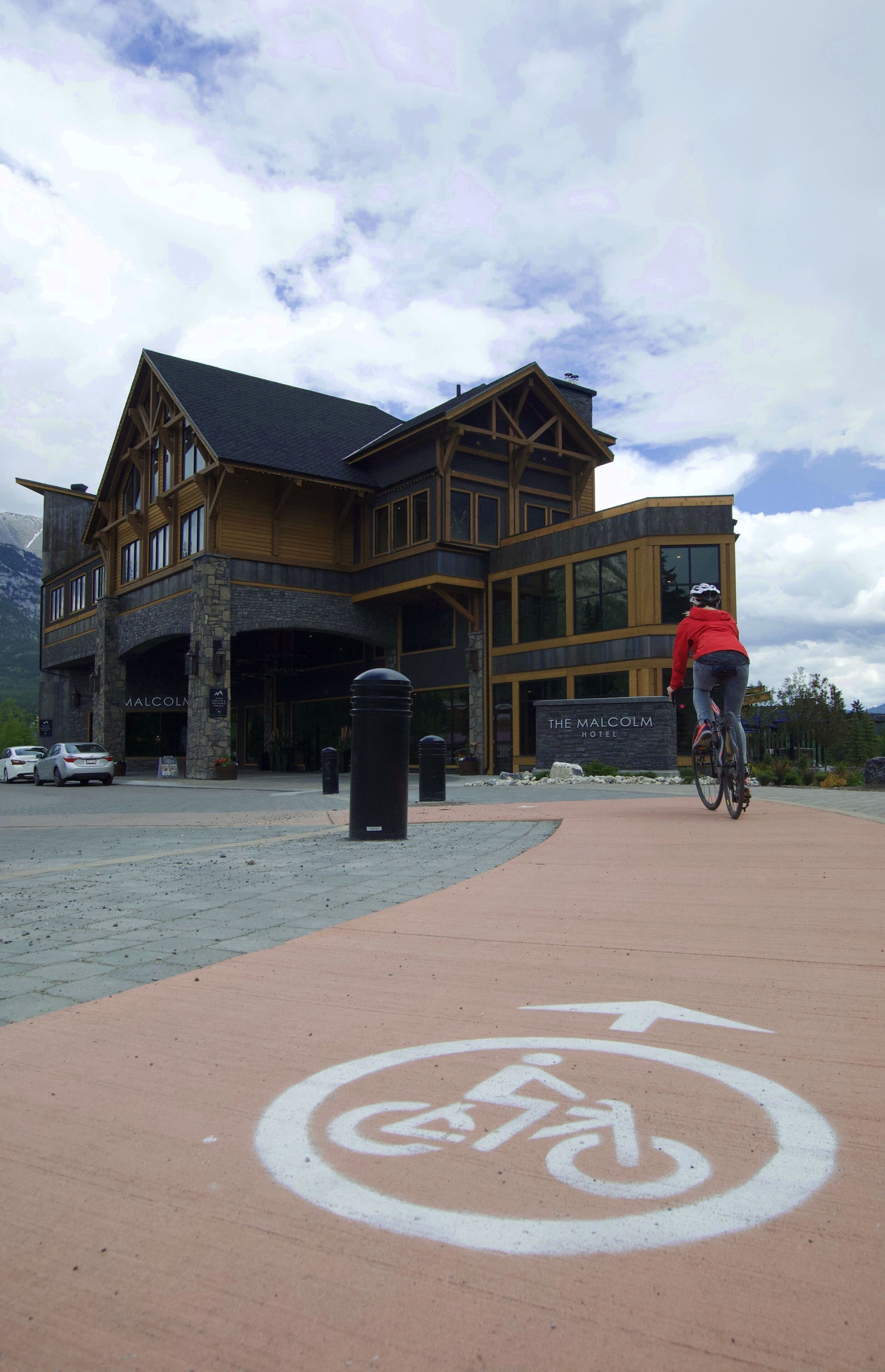 A hotel entrance with a bike path leading to it, a sign reading "The Malcolm Hotel", and a mountain backdrop.