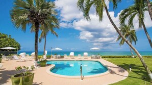 Zona de piscina con sillones de playa y sombrillas, rodeada de palmeras y vista al océano.