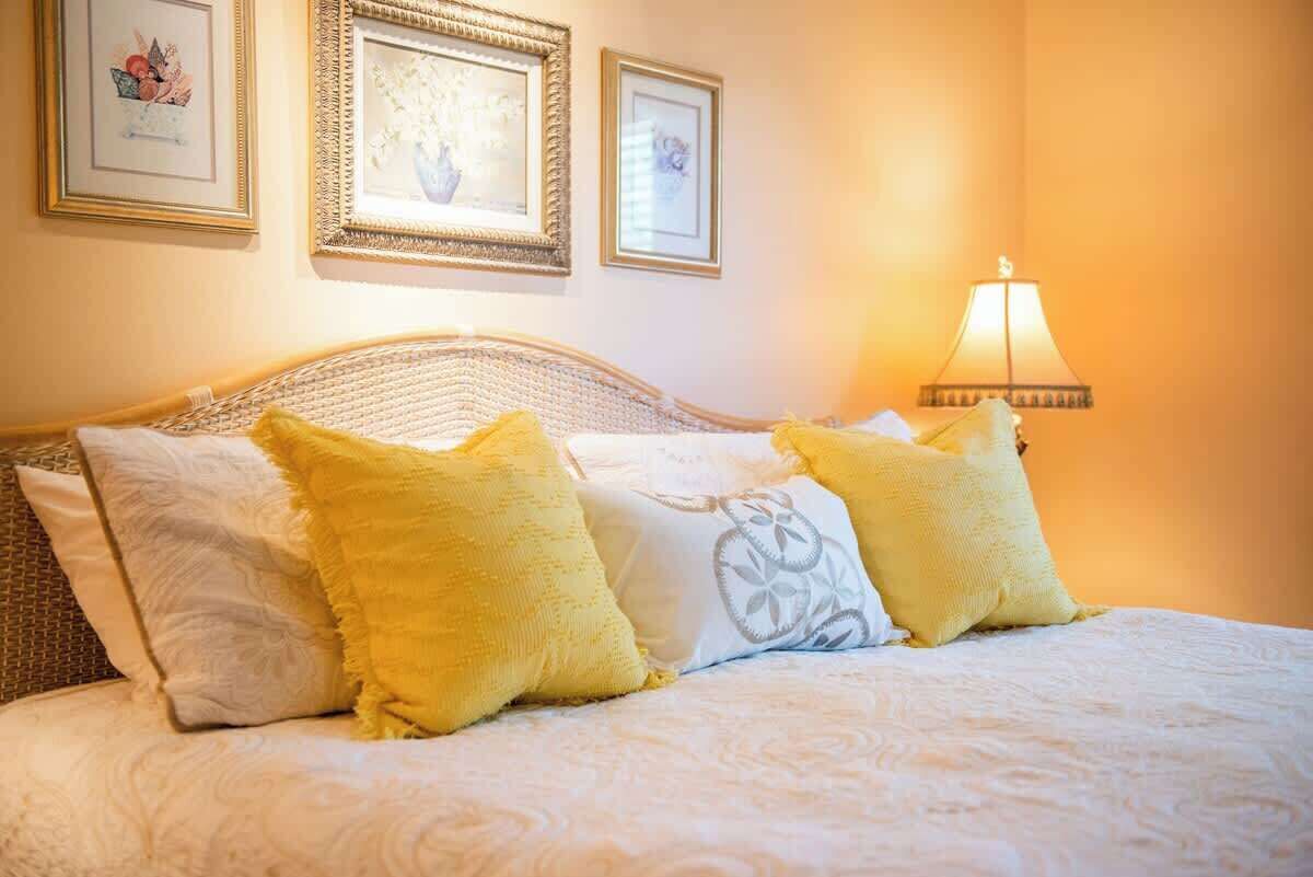 Ein ordentlich bezogenes Bett mit weißen und gelben Kissen, einem gewobenen Kopfteil und einer an der Wand befestigten Lampe.