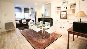 A modern living room with a glass dining table, white chairs, a sofa, and a fireplace. - Capitol Hill 2 Bedroom, 6ppl, Patio, Free Parking, near Capitol, Eastern Market (Washington)