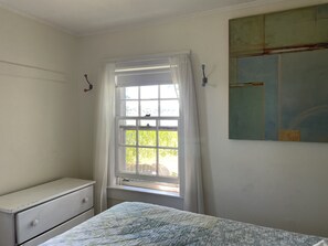 Een slaapkamer met een enkel bed, een raam met gordijnen, een klein witte dressoir en een schilderij aan de muur.