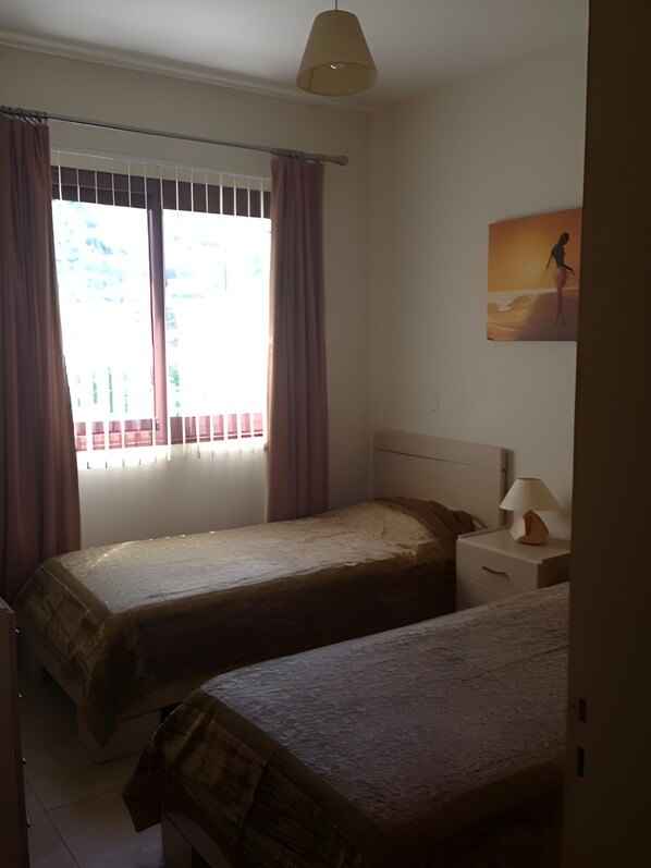 Un dormitorio con dos camas individuales, una mesita de noche con lámpara, una ventana con cortinas y un cuadro en la pared.