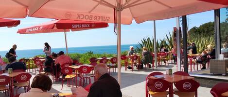 An outdoor seating area with red chairs and tables, umbrellas, and a view of the sea.