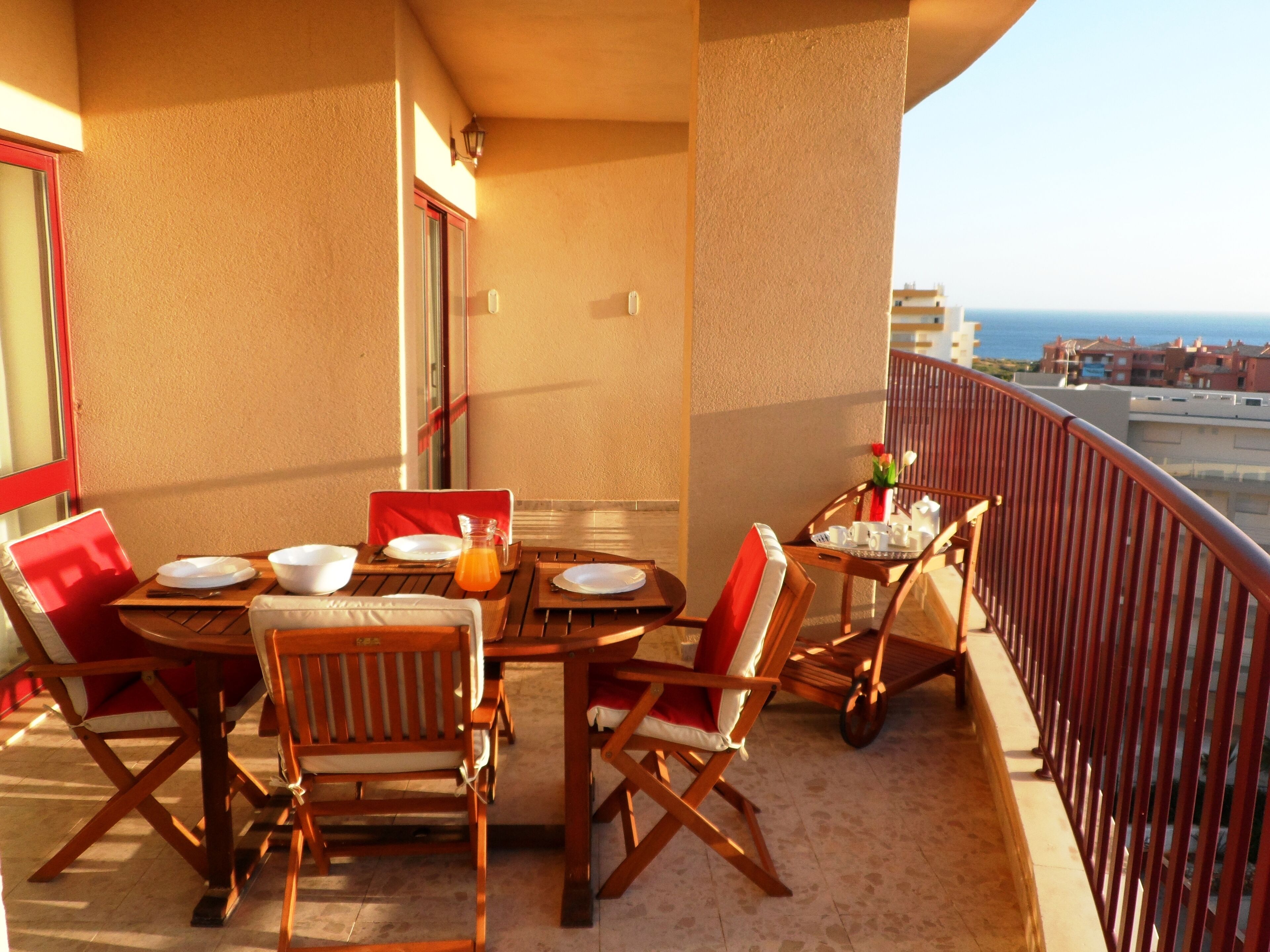Un balcón con una mesa preparada para dos, sillas, una mesita con una jarras de jugo de naranja y vista al mar.