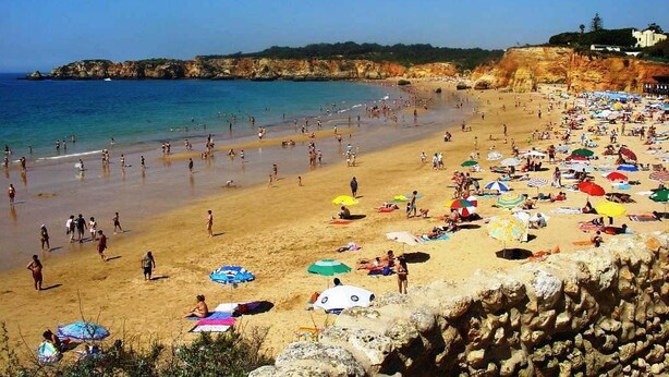 Una playa concurrida, gente tomando el sol y nadando, y un acantilado rocoso al fondo.