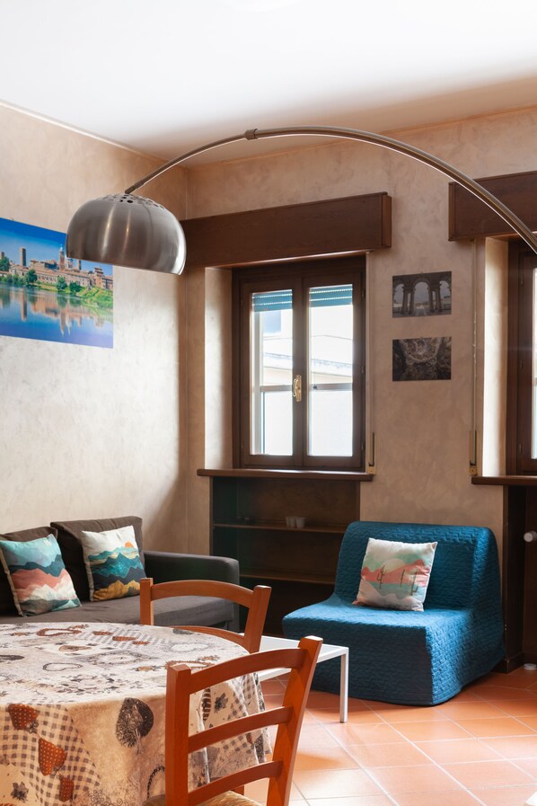 A cozy living room with a round table, chairs, a blue sofa, a large window, and a wall-mounted lamp.