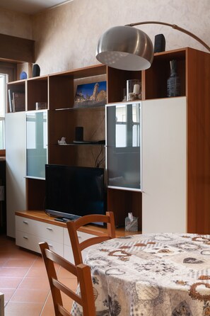 A room with a wooden TV stand, a flat-screen TV, a dining table with a patterned tablecloth, and a wooden chair.