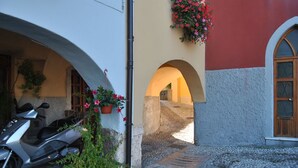 A narrow cobblestone street with a scooter parked on the left, a potted plant with flowers, and an arched doorway leading to a building with a red and white facade.