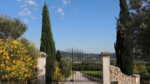 A gated entrance with a stone wall and a metal gate, surrounded by tall cypress trees and yellow flowering bushes. - DROME-LIMIT VAUCLUSE-GÎTE WITH SWIMMING POOL - PANORAMIC VIEW (Vinsobres)