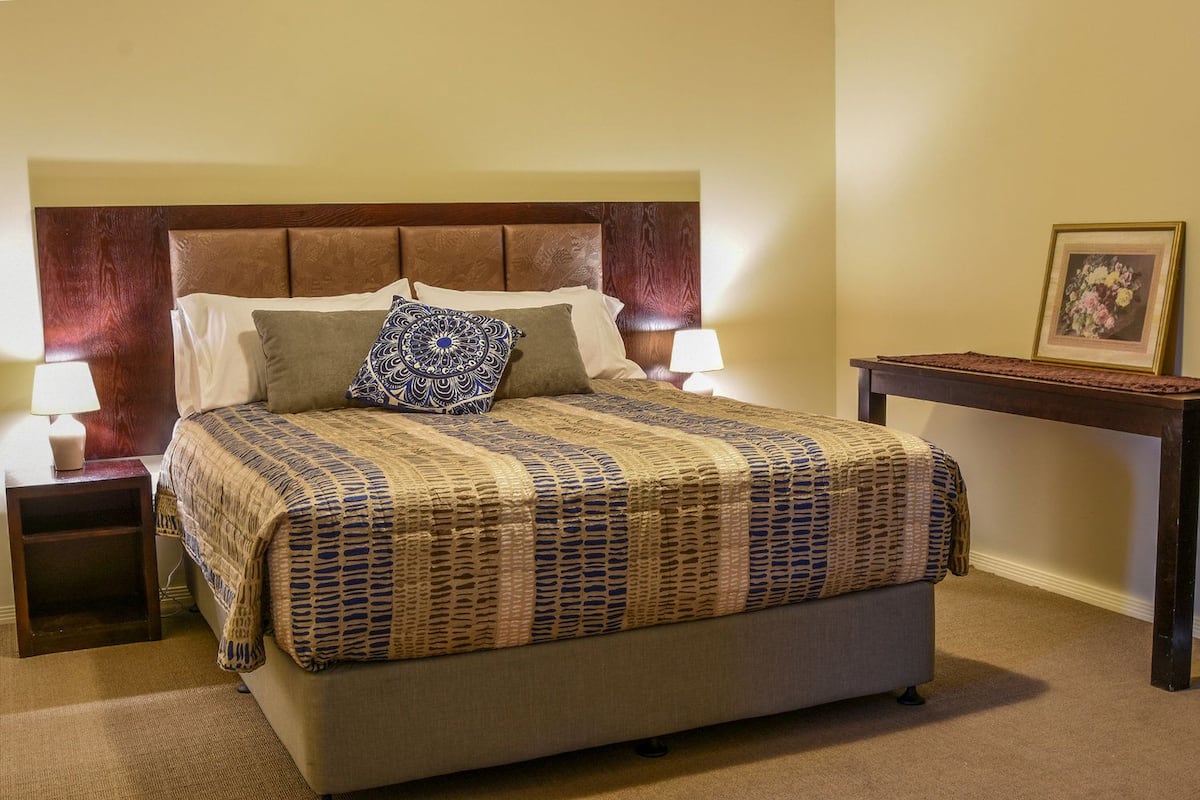 A neatly made bed with a patterned bedspread, two pillows, and a decorative pillow. There is a bedside table with a lamp and a framed picture on the wall.