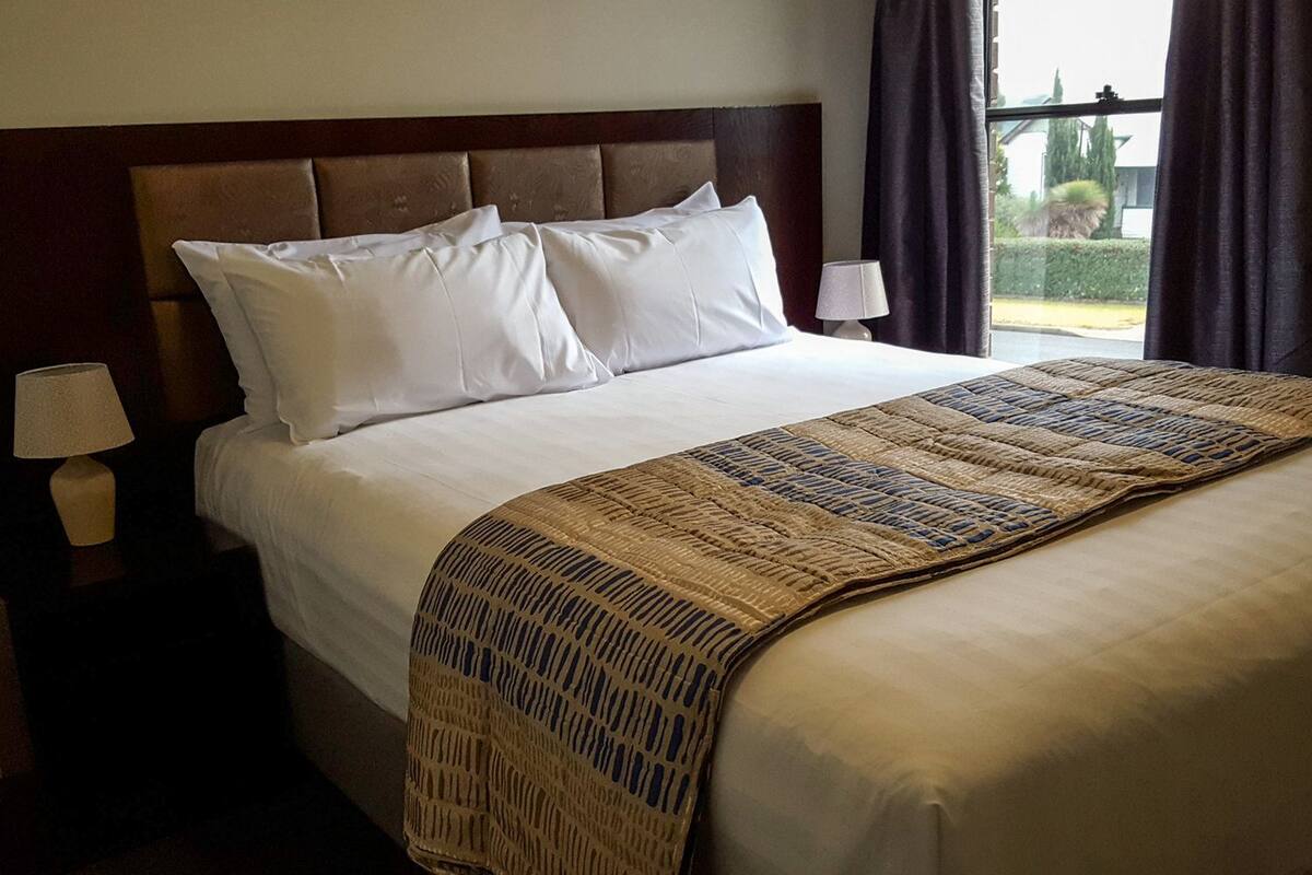 A neatly made bed with white linens, a patterned throw blanket, and a wooden headboard. There are two bedside lamps and a window with curtains.