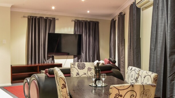 A living room with a dark wooden table, patterned armchairs, a sofa, and a flat-screen TV mounted on the wall.