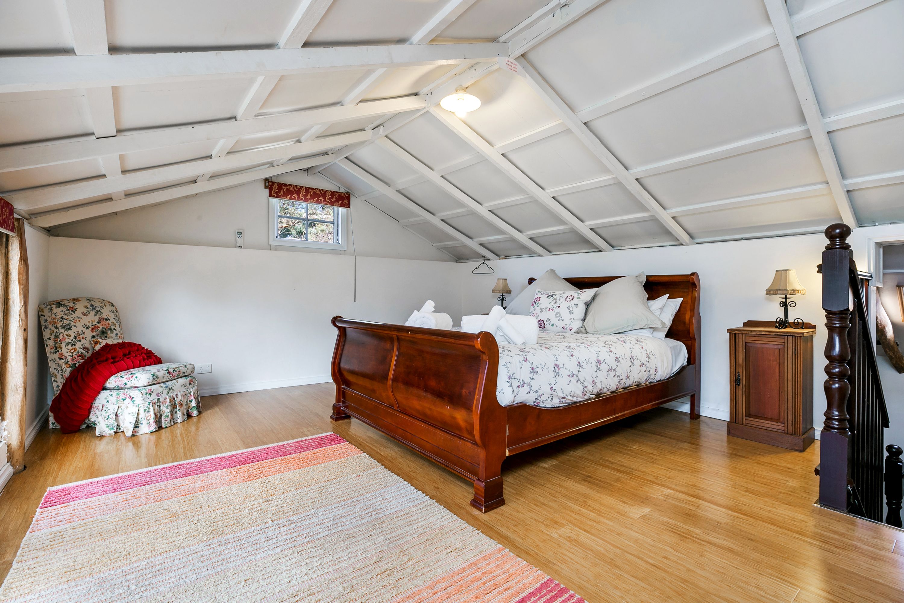 A bedroom with a wooden bed, a small stool, a red cushion, a nightstand, and a lamp.
