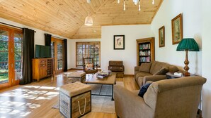 Amplia sala de estar con piso de madera, techo alto y ventanas grandes que dejan entrar luz natural. Hay un sofá, una mesa de centro y una estantería.