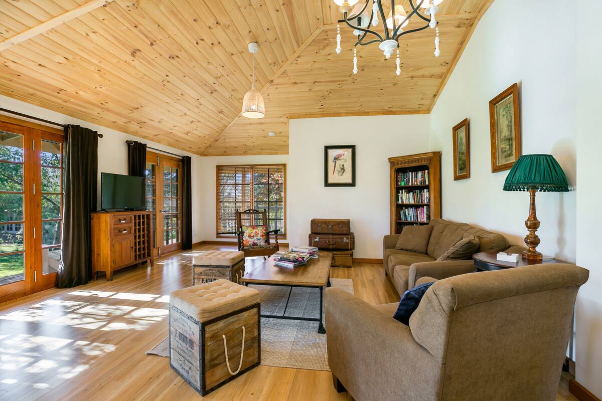 Amplia sala de estar con piso de madera, techo alto y ventanas grandes que dejan entrar luz natural. Hay un sofá, una mesa de centro y una estantería.